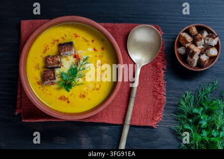 Oberblick auf gelbe Kürbiskremesuppe mit Croutons. Vegetarische Linsensuppe oder Kürbis-Püreesuppe garniert mit Croutons, Paprika und frischen Kräutern in Tontopf Stockfoto
