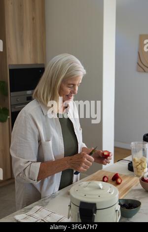 Seniorin schneidet Erdbeeren auf einem Schneidebrett in der Küche, macht einen Smoothie, trägt ein helles Hemd Stockfoto