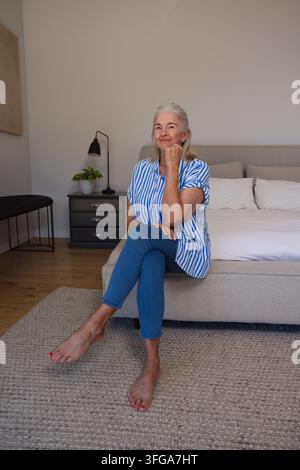 Seniorfrau sitzt auf Bett im Schlafzimmer, ruht das Kinn auf der Hand, trägt gestreifte Bluse und Jeans Stockfoto