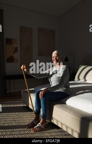 Seniorin sitzt auf Bettkante, hält hölzernen Stock und lächelt in Richtung Sonnenlicht im Schlafzimmer Stockfoto
