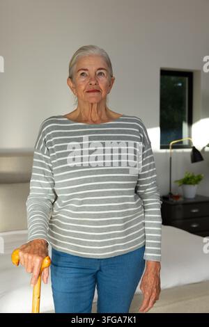 Seniorin steht im Schlafzimmer am Bett, trägt gestreiftes Hemd und Jeans, hält einen Holzstock Stockfoto