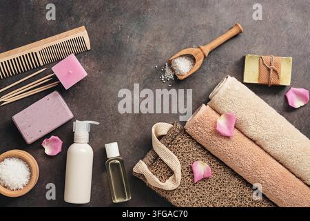 Flat Lay ein Set aus verschiedenen Badezimmeraccessoires. Frottierhandtuch, Seife, Kamm, Öl, Shampoo, Luffa-Waschhandschuh, Meersalz auf einem dunklen rustikalen Hintergrund. Blick von oben Stockfoto