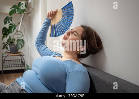 Erschöpfte übergewichtige junge Frau auf dem Sofa, die unter Überhitzung im Innenraum leidet und einen Handfächer benutzt, um sich abzukühlen, schwer atmend. Stickige Luft, Unbehagen, fehlende Klimaanlage, hohe Temperatur zu Hause. Stockfoto