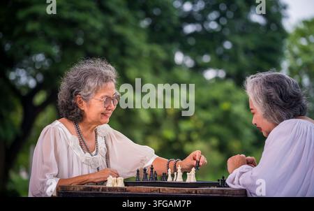 Ältere Frauen sitzen an einem Holztisch im Freien und spielen Schach, wobei sie eine Schachfigur halten. Stockfoto