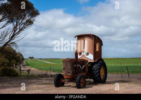 Ein rostiger Vintage-Traktor, der mit einer überdimensionalen, maßgefertigten Kabine mit bemalten Cartoon-Augen modifiziert wurde, steht auf einem Schotterweg neben grünen landwirtschaftlichen Feldern Stockfoto