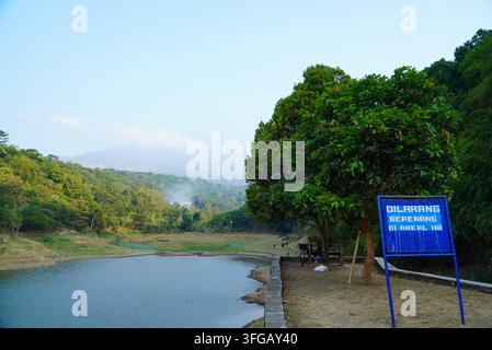 Ruhiger See spiegelt entfernte Bäume und neblige Hügel wider. Blaues Schild steht nahe dem Ufer und markiert einen ruhigen Ort. Stockfoto