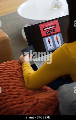 Frau sitzt auf der Couch zu Hause, hält eine Kreditkarte und durchsucht einen Online-Shop auf dem Laptop Stockfoto