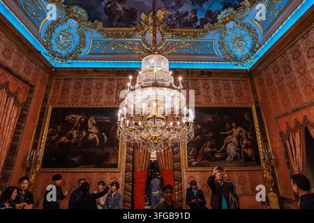 Madrid, Spanien: 7. Apr. 2025 – Eine Menschenmenge steht unter einem großen vergoldeten Kronleuchter in einem eleganten, mit Damast tapezierten Raum, an dessen Wänden Porträtgemälde hängen Stockfoto