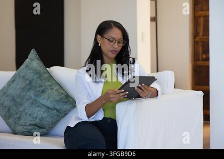 Afroamerikanische Frau sitzt auf dem Sofa zu Hause, trägt ein grünes Oberteil, eine Brille und hält ein Tablet Stockfoto