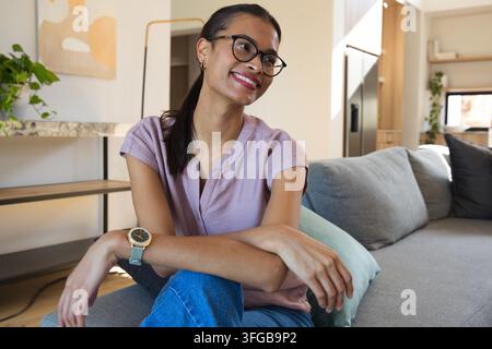 Afroamerikanische Frau sitzt auf grauem Sofa im Wohnzimmer, trägt malvenfarbene Bluse und Brille Stockfoto