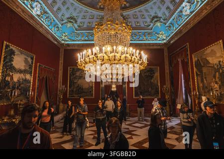 Madrid, Spanien: 7. Apr. 2025 – Eine Menschenmenge steht unter einem großen vergoldeten Kronleuchter in einem eleganten, mit Damast tapezierten Raum, an dessen Wänden Porträtgemälde hängen Stockfoto