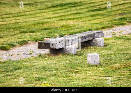 Eine hölzerne Parkbank steht auf einem üppigen grünen Rasen neben einem Kopfsteinpflasterweg in einer ruhigen Gedenkstätte unter sanftem Tageslicht. Die Szene Stockfoto