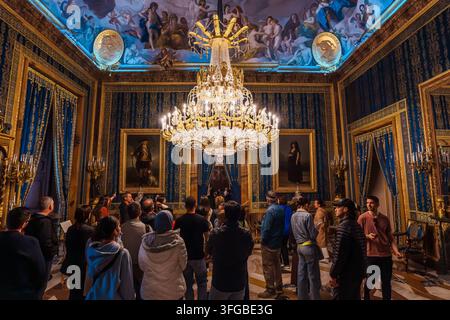 Madrid, Spanien: 7. Apr. 2025 – Eine Menschenmenge steht unter einem großen vergoldeten Kronleuchter in einem eleganten, mit Damast tapezierten Raum, an dessen Wänden Porträtgemälde hängen Stockfoto