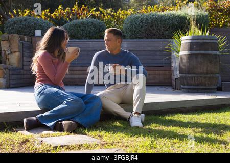 Verschiedenes Paar sitzt auf einer erhöhten Holzterrasse im Garten und hält Keramikbecher, trägt Pullover Stockfoto