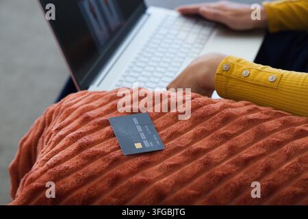 Afroamerikanische Frau tippt auf einem Laptop zu Hause, Kreditkarte auf orangefarbenem Kissen, gelber Ärmel Stockfoto