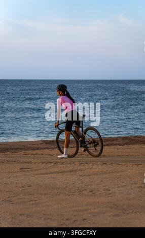 Eine junge Radfahrerin in einem pinkfarbenen Trikot, die auf ihrem Rennrad sitzt und bei Sonnenaufgang auf einem Sandstrand auf das Mittelmeer blickt. Stockfoto