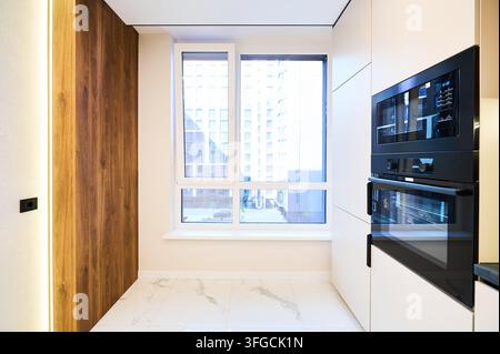 Integrierter Ofen und Mikrowelle neben großem Fenster in modernem Kücheninterieur. Helle Wohnraumfläche mit weißen Schränken, Holzakzentwand und minimalistischem Premium-Designkonzept Stockfoto