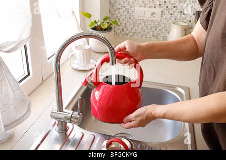 Frauenhände füllen einen roten Wasserkocher mit Leitungswasser im Küchenwaschbecken. Stockfoto