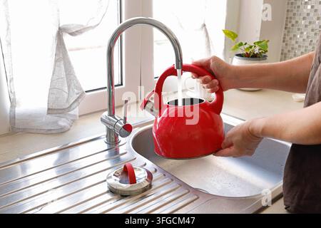 Frauenhände füllen einen roten Wasserkocher mit Leitungswasser im Küchenwaschbecken. Stockfoto