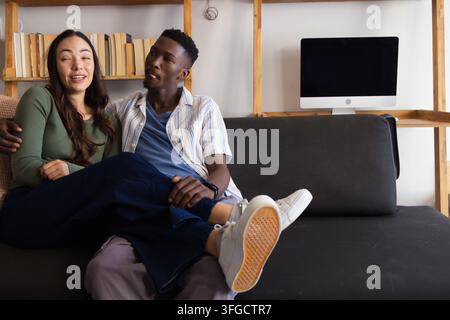 Paar sitzt auf dunklem grauem Sofa zu Hause, Frau legt ihre Beine über den Schoß des Mannes, Bücherregal sichtbar Stockfoto