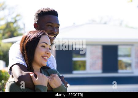 Verschiedenes Paar steht im Vorgarten, Mann umarmt Frau, lächelt, blickt in die Kamera, Haus im Hintergrund Stockfoto