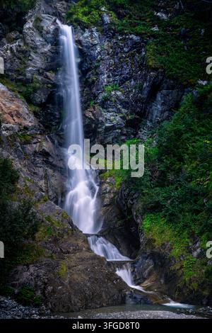 Langzeitvertikalaufnahme eines wunderschönen alpinen Wasserfalls, der über dunkle Felsen stürzt und von üppiger grüner Vegetation in Feichten im Kaunertal umgeben ist Stockfoto