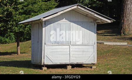 Kleiner geschlossener weißer Holzmarktkiosk, der auf Betonblöcken auf einem grasbewachsenen Rasen in einer sonnigen Parkumgebung steht, umgeben von grünen Bäumen Stockfoto