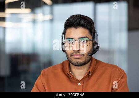 Nahaufnahme eines konzentrierten Mannes mit Brille und Headset in einem modernen Büro mit weichem unscharfem Hintergrund, der direkt in die Kamera schaut für Remote-Support, Beratung und Kundenservice Stockfoto