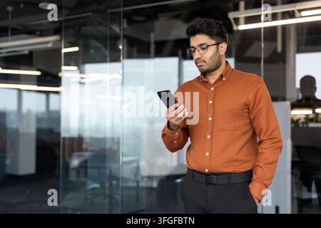 Porträt eines jungen Berufstätigen in Geschäftskleidung, der in einem zeitgenössischen Glasbüro steht und ruhig sein Smartphone in der Nähe einer Glaswand mit subtilen Reflexionen und sanfter Innenbeleuchtung überprüft Stockfoto