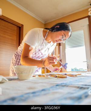 Junge Frau in einer Schürze, die selbstgemachte Kekse mit Zuckerguss an einem Küchentisch dekoriert und eine warme kreative Backaktivität zu Hause genießt. Stockfoto