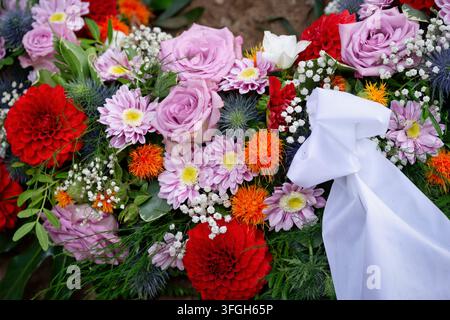 Nahaufnahme eines bunten Trauerkranzes mit lila Rosen, roten Dahlien und rosa Chrysanthemen mit einem eleganten weißen Satinband. Stockfoto