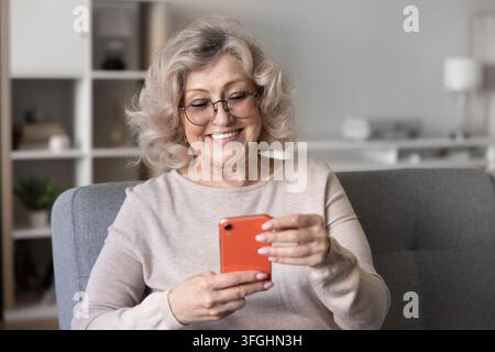 Lächelnde Seniorin überprüft Smartphone, sitzt auf der Couch zu Hause Stockfoto