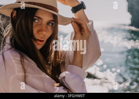 Frauen-Ozean-Porträt junge Frau in weißem Leinenhemd, die ihren Strohhut am Meer während der goldenen Stunde bei Sonnenuntergang anpasst Stockfoto