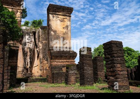 Reihen von Laterit-Säulen neben einer verwitterten Buddha-Statue und einem Backsteinturm im historischen Park Si Satchanalai, Sukhothai, Thailand Stockfoto