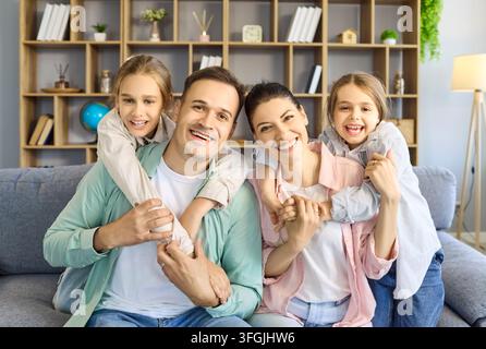 Zufriedene Eltern mit zwei Töchtern, die sich zu Hause auf dem Sofa umarmen, lächelndes Innenporträt Stockfoto