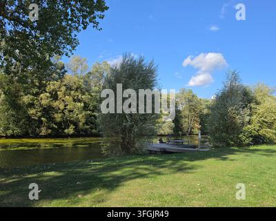 Scenische Ansicht eines ruhigen Flusses, der durch einen grünen Park mit üppigen Bäumen und einer kleinen hölzernen Pierbrücke unter einem strahlend blauen Himmel fließt Stockfoto
