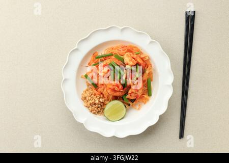 Frische Pad-Thai-Nudeln in einer weißen muschelförmigen Schüssel. Schwarze Essstäbchen liegen neben dem Gericht. Garniert mit grünen Zwiebeln, zerkleinerten Erdnüssen und Limette Stockfoto