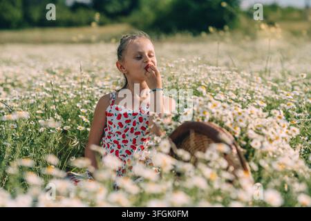 In einem Gänseblümchenfeld isst ein Mädchen in einem Kirschkleid eine Erdbeere. Ihre Augen sind geschlossen. Ein Korb steht neben ihr. Stockfoto