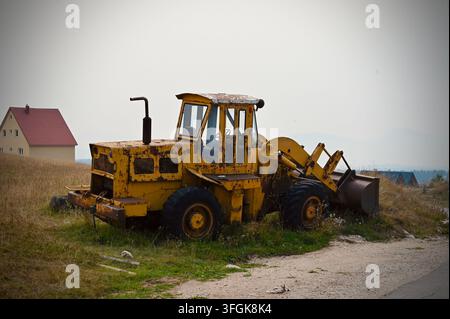 Nahaufnahme Seitenansicht eines rostigen, verwitterten gelben Radladers, das auf einem grasbewachsenen Feld neben einer Schotterstraße geparkt ist, mit einem kleinen Haus im Stockfoto