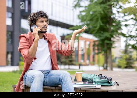 Verärgerter männlicher Student, der draußen ein Telefonat über ein Universitätsgruppenprojekt führt. Stockfoto