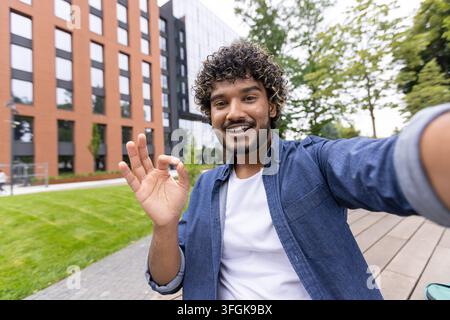 Junger Student nimmt ein Video-Selfie mit dem Smartphone auf und zeigt ein Ok-Zeichen auf dem Universitätsgelände. Stockfoto