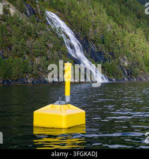 Blick auf eine hellgelbe Navigationsboje, die im ruhigen Wasser vor einem kaskadierenden Wasserfall an einer bewaldeten Klippenküste in Liabygda, More og Roms liegt Stockfoto
