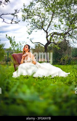 Junge Frau in einem weißen Hochzeitskleid lehnt auf einem vintage Ledersessel in einer üppigen grünen Wiese unter einem Baum Stockfoto