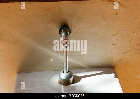 Ein Chrom-Wasserhahn, der an einer strukturierten Putzwand über einem weißen Waschbecken montiert ist, wird von warmem Sonnenlicht beleuchtet und wirft einen scharfen Schatten in einer rustikalen Inneneinrichtung Stockfoto