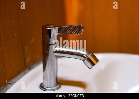 Ein chromfarbener Badezimmer-Wasserhahn mit strukturiertem Hebel, montiert auf einem weißen Keramikwaschbecken vor einer warmen Holzwand, saubere Sanitärarmatur-Details, die modernes Interieur schaffen Stockfoto