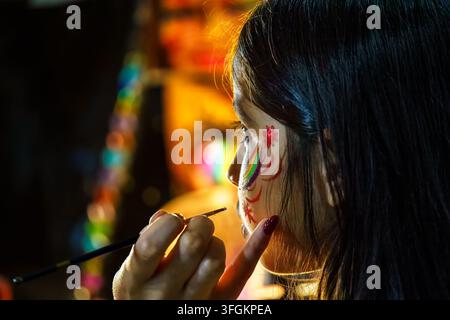 Weiches Licht erhellt ihr Gesicht. Bunte Farbe ziert ihre Wange mit sanfter Sorgfalt. Ihr Blick zeigt ruhige Konzentration. Der Pinsel taucht in lebhafte Farbtöne. Perfe Stockfoto