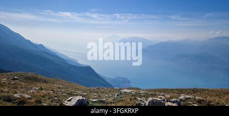 Scenische ultrabreite Landschaft des nebligen blauen Gardasee-Beckens, gesehen über einen grasbewachsenen Kamm unter einem hellen Herbsthimmel - Gardasee vom Monte Baldo-Kamm aus Stockfoto