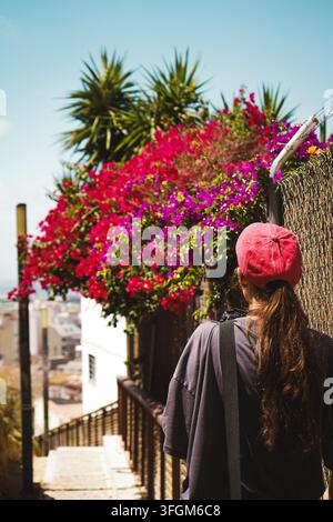 Rückansicht einer Frau, die einen sonnigen Hügelpfad entlanggeht, umgeben von farbenfroher Bougainvillea und mediterraner Vegetation in Alicante, Spanien, im Juni Stockfoto
