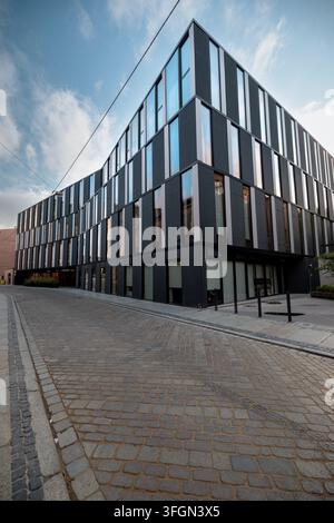 Gekrümmtes zeitgenössisches Bürogebäude mit abwechselnd dunkler Verkleidung und vertikalen Fenstern spannt sich über eine schmale Straße im Stadtzentrum. Stockfoto