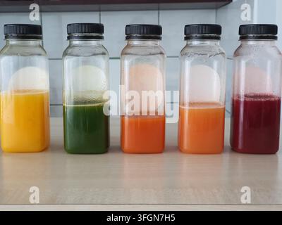 Fünf klare Glasflaschen mit schwarzen Schraubverschlüssen, die in einer Reihe auf einer hellen Holzarbeitsplatte angeordnet sind, jede enthält frische Säfte in verschiedenen Farben. Stockfoto